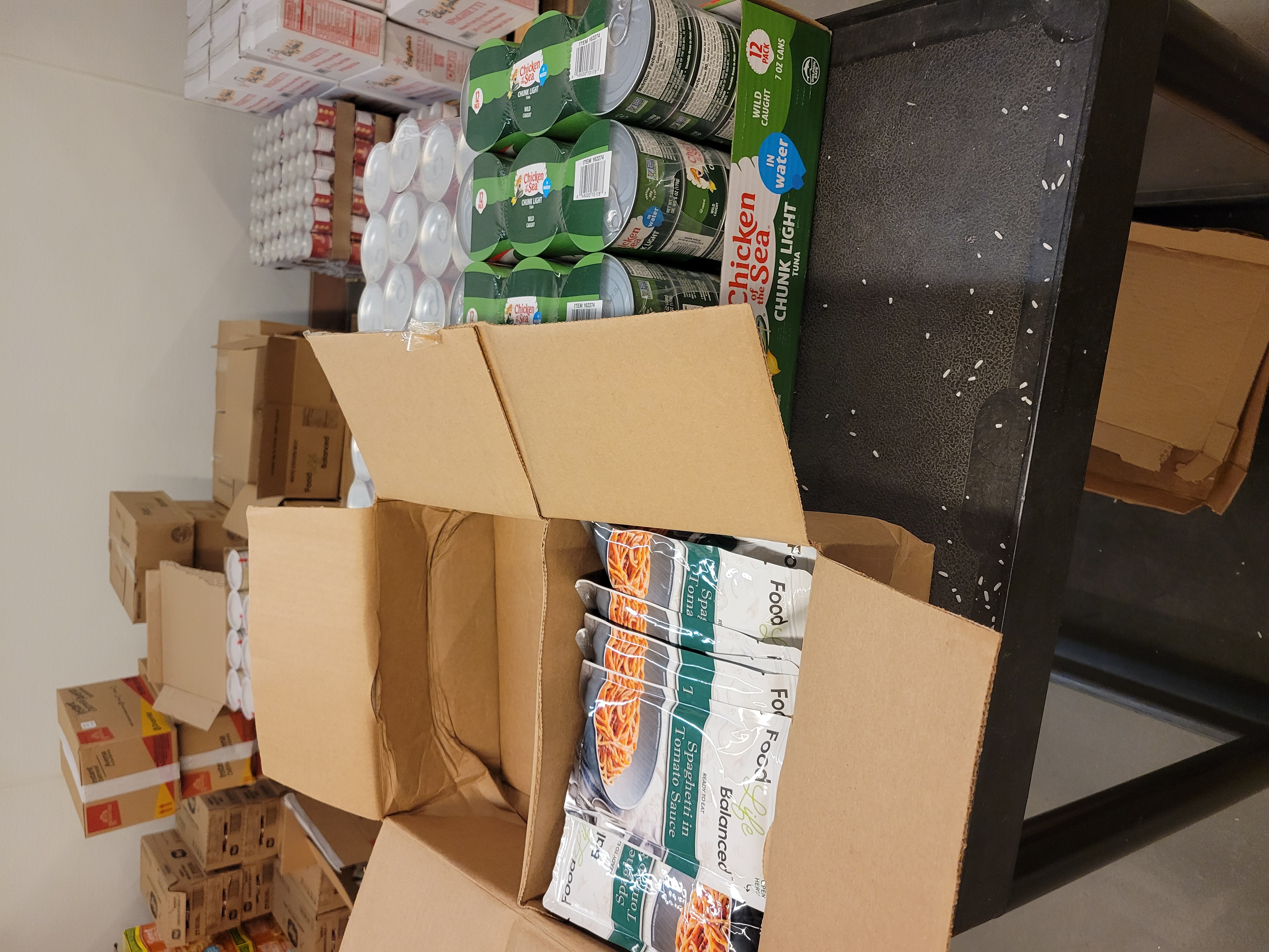 Box of canned tuna and box of spaghetti in tomato sauce pouches on top of a shelf cart with pallets of food behind it.